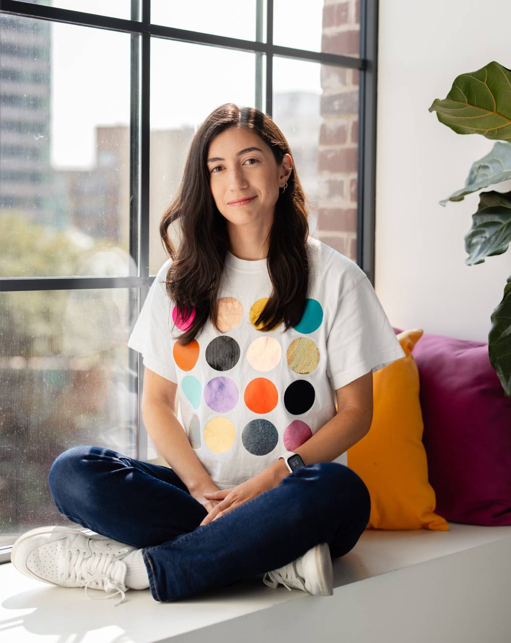 A 30-something smiling brunette white woman, wearing a multicoloured polka dot shirt, dark blue jeans, and white sneakers, sitting cross legged in front of a window